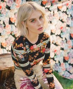 a woman sitting on top of a wooden bench in front of a flower covered wall