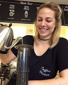 a woman pours coffee into a cup from a stainless steel kettle while smiling at the camera