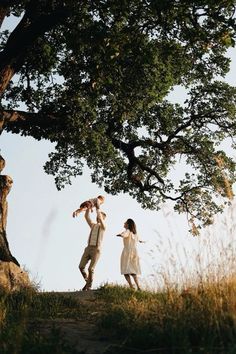 a man and woman standing under a tree on top of a grass covered hill next to a forest