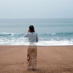 a woman is standing on the beach talking on her cell phone and looking out at the ocean