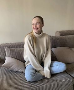 a young woman sitting on top of a couch wearing blue jeans and a turtle neck sweater