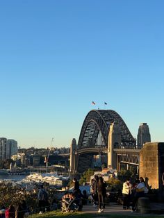 people are sitting on benches near the water in front of a bridge and some buildings