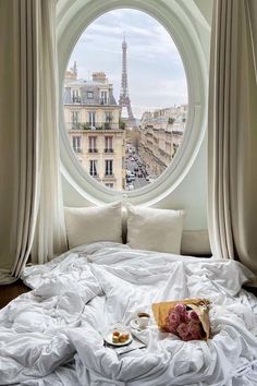 a bed with white sheets and pillows in front of a round window overlooking the eiffel tower