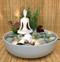 a white buddha statue sitting in a bowl filled with rocks and plants