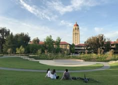 three people sitting on the grass in front of a clock tower