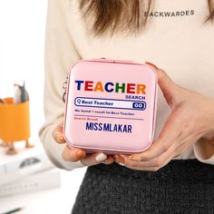 a woman holding a pink lunchbox with the words teacher on it in front of her