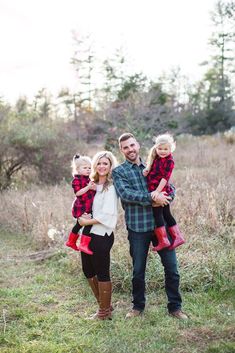 a man and woman holding two small children while standing in the grass with trees behind them