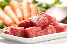 some raw meat on a white plate next to other fresh vegetables and greens in the background