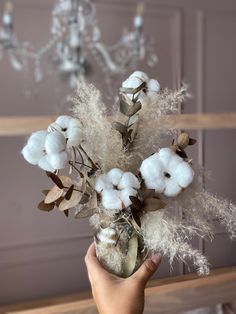 a hand holding a vase filled with cotton and other flowers in front of a chandelier