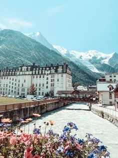 a river running through a lush green hillside next to tall buildings and snow covered mountains