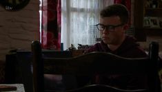 a man sitting at a table in front of a window with curtains on the windowsill