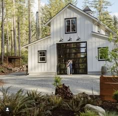 two people standing in front of a white barn with black doors and windows, surrounded by pine trees