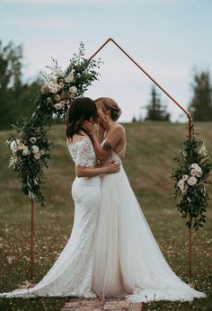 two women in wedding gowns kissing each other under an arch decorated with flowers and greenery