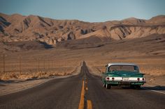 an old green car is driving down the road in front of some mountains and hills