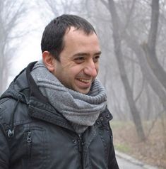 a man with a scarf around his neck smiles at the camera while standing in front of trees