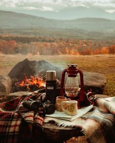 a picnic table with coffee cups and camera on it in front of a campfire