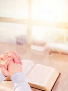 two hands clasped over an open book on a table with sunlight streaming through the window