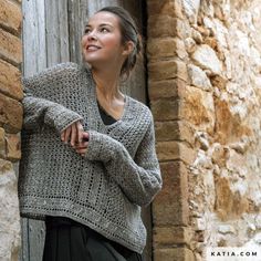 a woman standing next to a brick building wearing a gray crocheted sweater and black pants