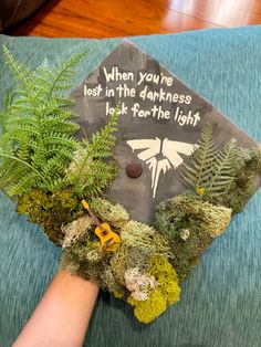 someones hand holding a graduation cap decorated with moss and fern leaves that reads, when you're lost in the darkness look for the light