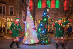 three men dressed in green and red standing next to a christmas tree with lights on it