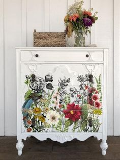 a white cabinet with flowers painted on the front and bottom, sitting against a wall