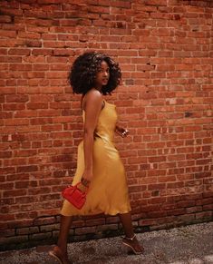 a woman in a yellow dress is walking by a brick wall holding a red purse