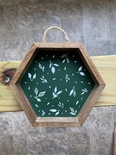 a hand holding a wooden frame with leaves painted on the inside and outside, in front of a tiled wall