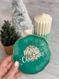 a hand holding a green plate with merry christmas written on it next to a potted plant