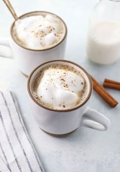 two mugs filled with hot chocolate and cinnamon on top of a white tablecloth