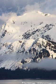 snow covered mountains rise above the water on a cloudy day with clouds in the foreground