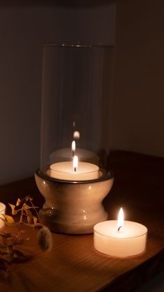 three lit candles sitting on top of a wooden table next to a glass container filled with leaves