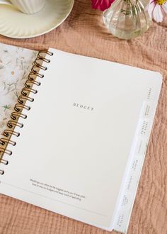 an open notebook sitting on top of a table next to some plates and flowers in vases