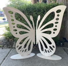 two white metal butterfly sculptures sitting on top of a cement ground next to a bush