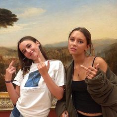 two young women standing next to each other in front of a wall with a painting on it