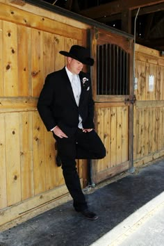 a man in a black suit and hat leaning against a wooden fence with his hands on his hips