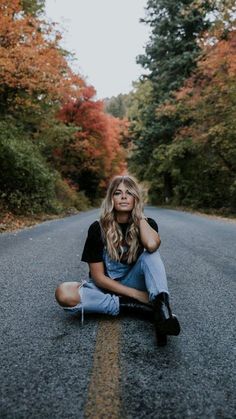 a woman sitting on the side of a road in front of trees with orange leaves