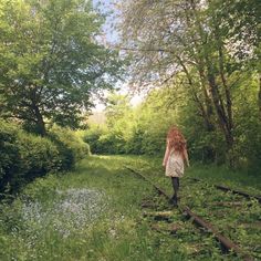 a girl walking on train tracks in the middle of some trees and grass, with her back to the camera