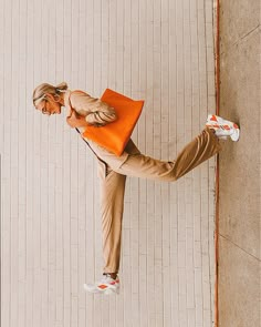 a woman standing on one leg and holding an orange bag in her other hand while leaning against a wall