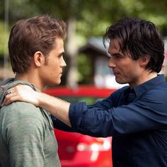 two young men standing next to each other in front of a red car, one holding his arm around the other's shoulder