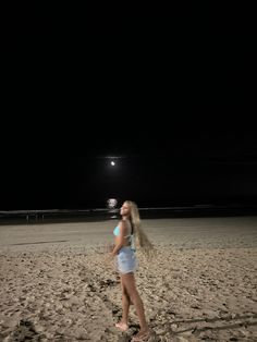 a woman standing on top of a sandy beach under a light at night with her hair blowing in the wind