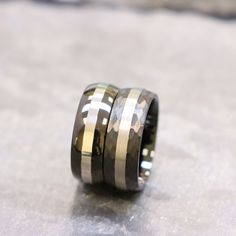 two wedding bands with gold and black inlays on each side, set against a stone surface