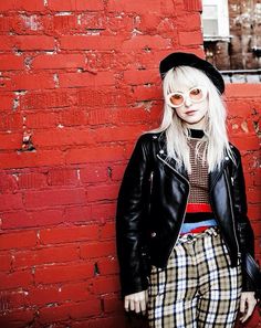 a woman with white hair and glasses standing in front of a red brick wall wearing plaid pants