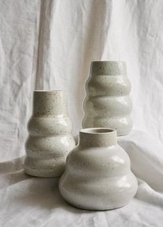 three white vases sitting next to each other on top of a white cloth covered surface