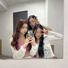 three young women are looking at their cell phones in front of a mirror, with one holding her hair up