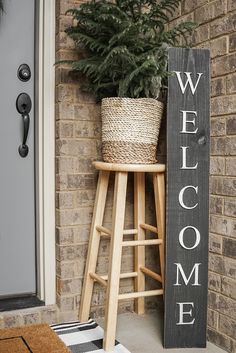 a welcome sign next to a wooden stool and potted plant on the front porch