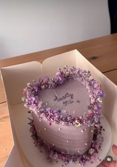 a heart shaped cake in a white box with purple flowers on it and someone's hand