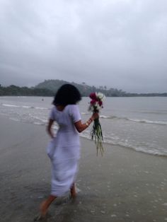 a woman is walking on the beach with flowers in her hand and water behind her