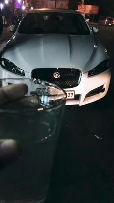 a hand holding a glass in front of a white car on the street at night