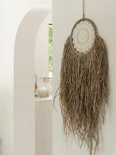 a white wall hanging with a large piece of grass on it's side and a vase in the background