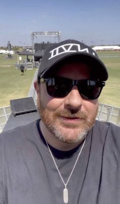 a man wearing sunglasses and a hat at a baseball game, taking a selfie